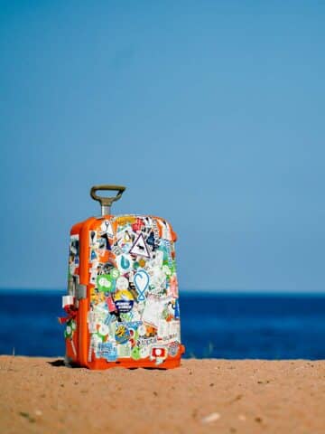 An orange suitcase with stickers all over it left on a sandy beach. The blue sea is visible in the background.