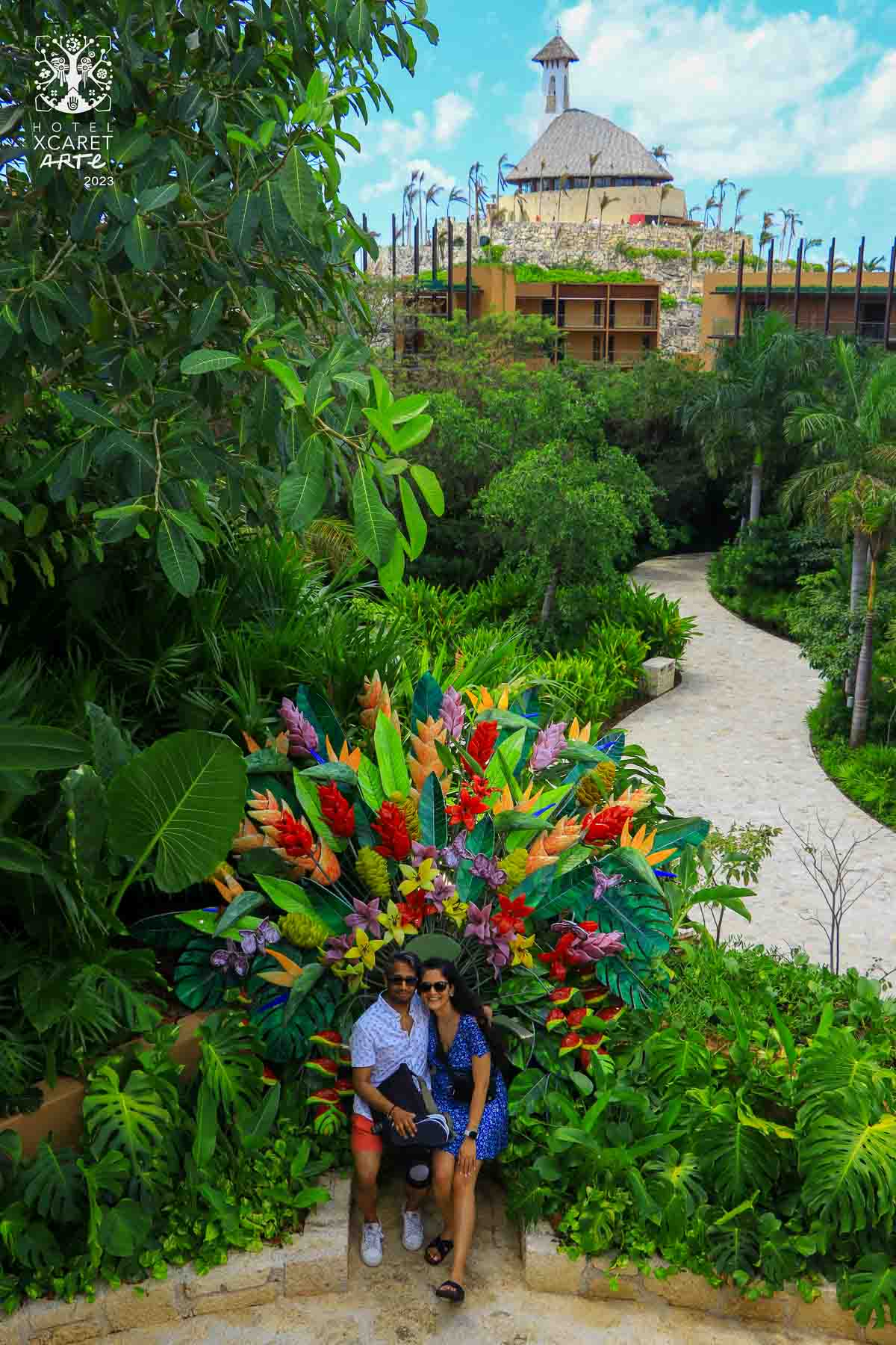 A xelfie taken in Xcaret Arte of Riz and Reem sitting in a chair surrounded by flowers and greenery. The pyramid can be seen in the background.