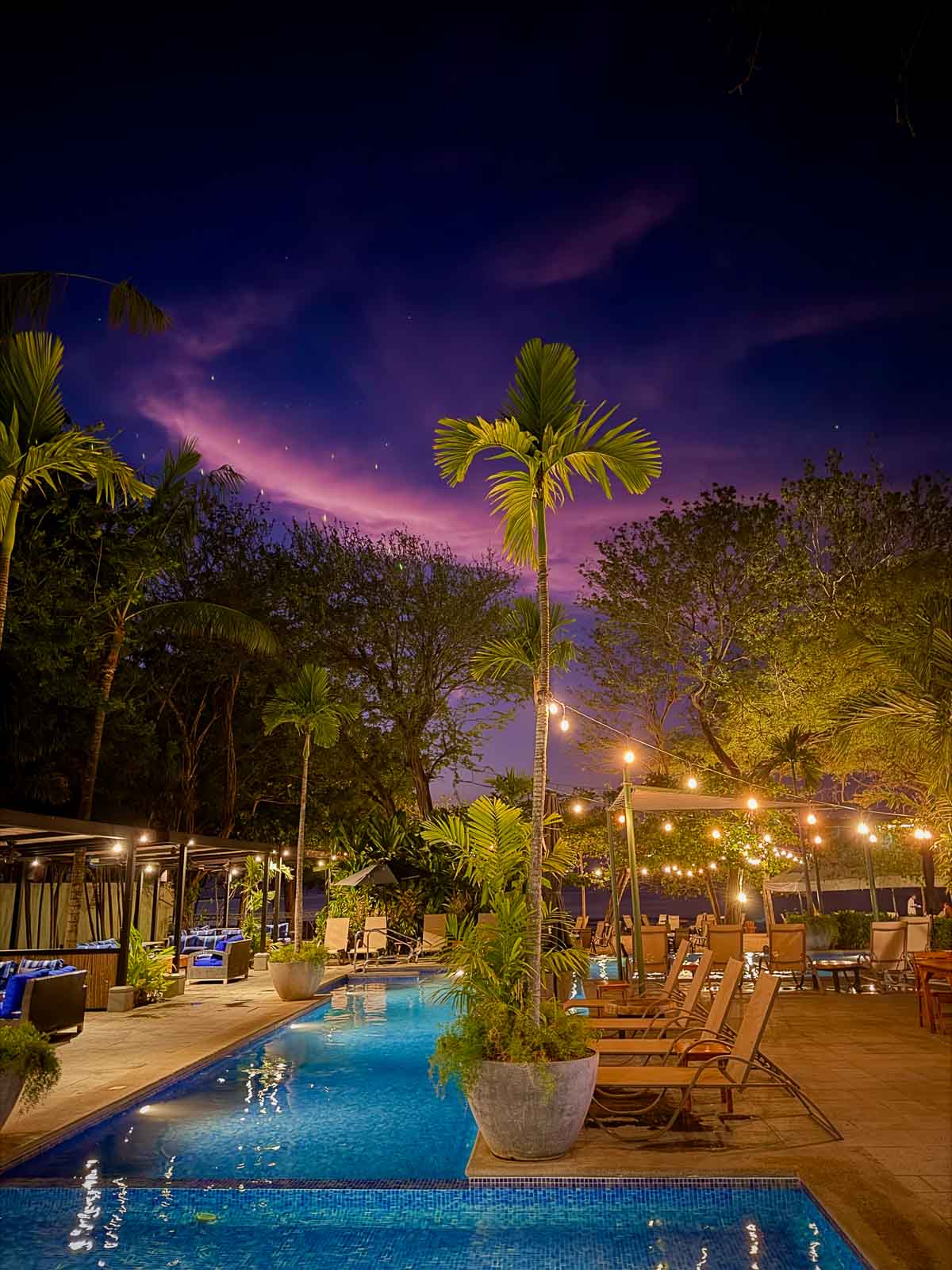 Langosta Beach Club at Night shows the blue pool, deck chairs, twinkle lights, and a dramatic cloud pattern with pink and purple hues.