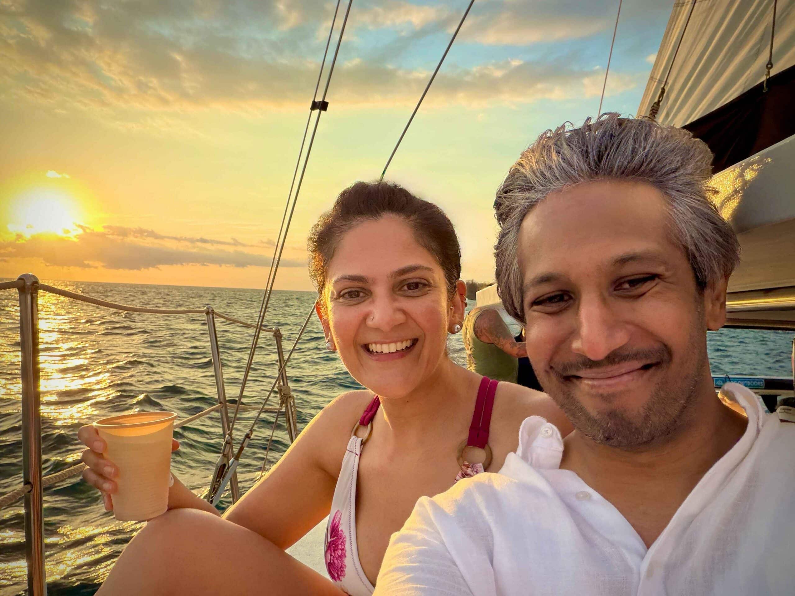 Riz and Reem sitting on a catamaran cruise at sunset. The yellow sun is dropping behind them towards the ocean waves with some dramatic cloud patterns.
