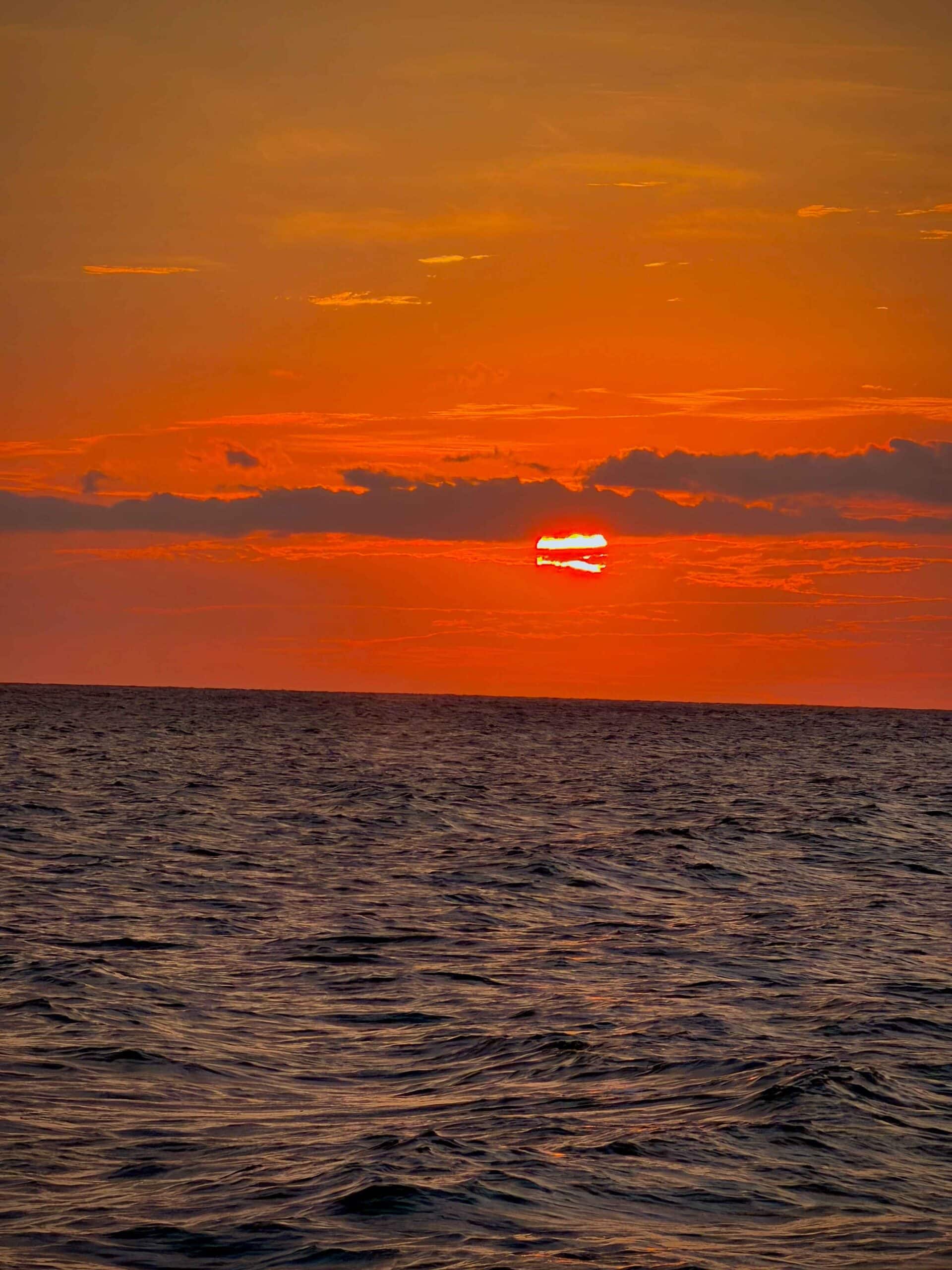 A dramatic shot of the red sunset, slightly obscured by crossing clouds, over the choppy waters of the Pacific Ocean.
