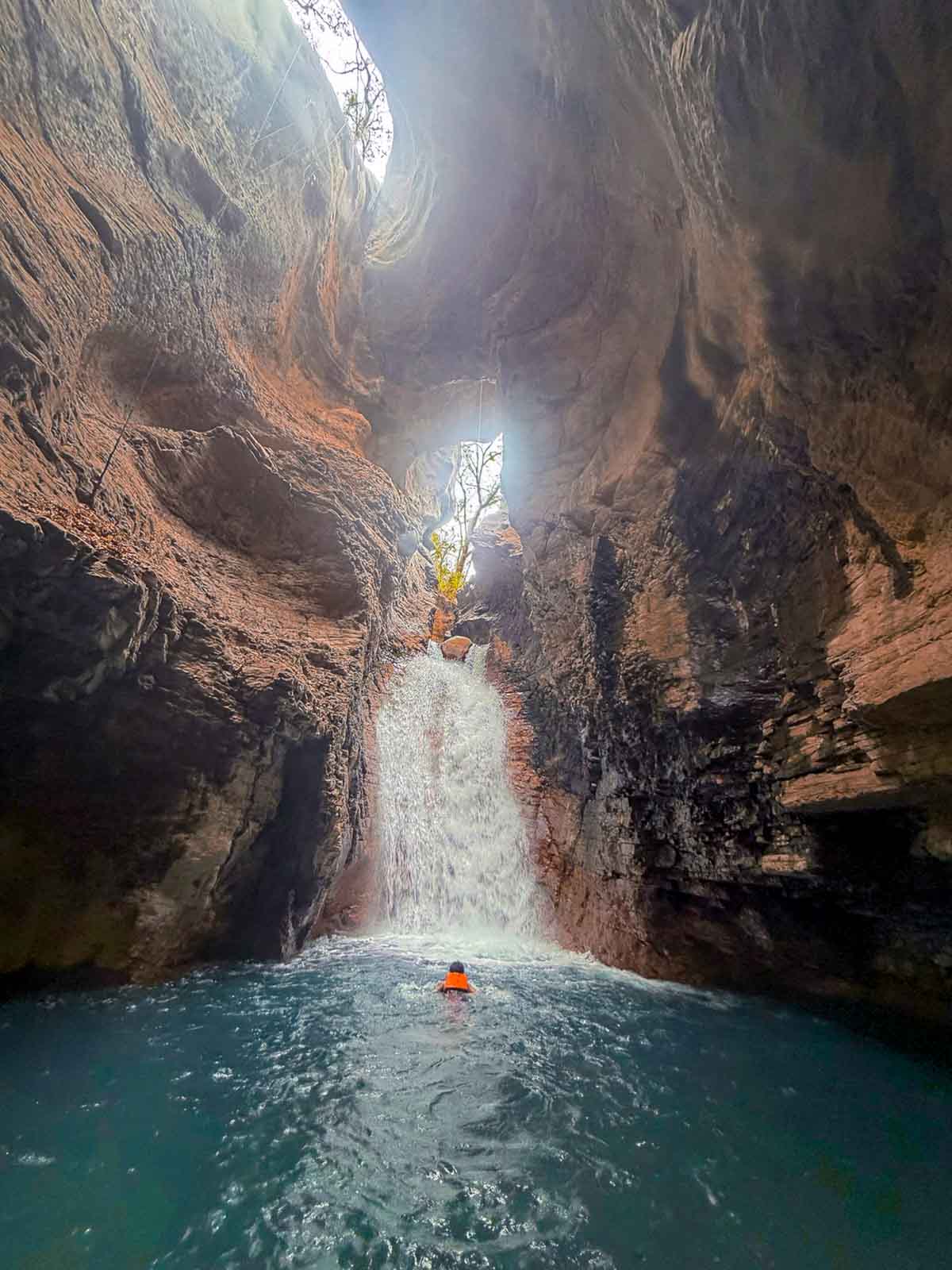 Riz swims up to the waterfall in La Leona Waterfall. The waterfall is enclosed by rock faces on both sides and light peeks through the opening where the water rushes in from.