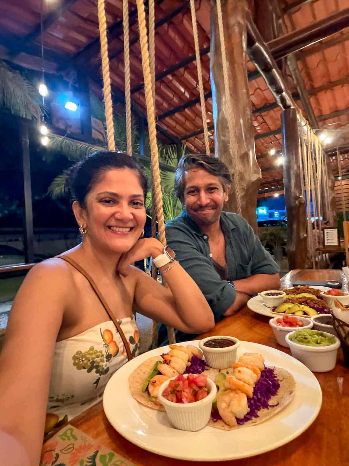 Reem and Riz sitting in the Green Papaya Taco restaurant (Tamarindo, Costa Rica) with tacos in front of them. They are both sitting on swings which are one of the more unique qualities of the restaurant.