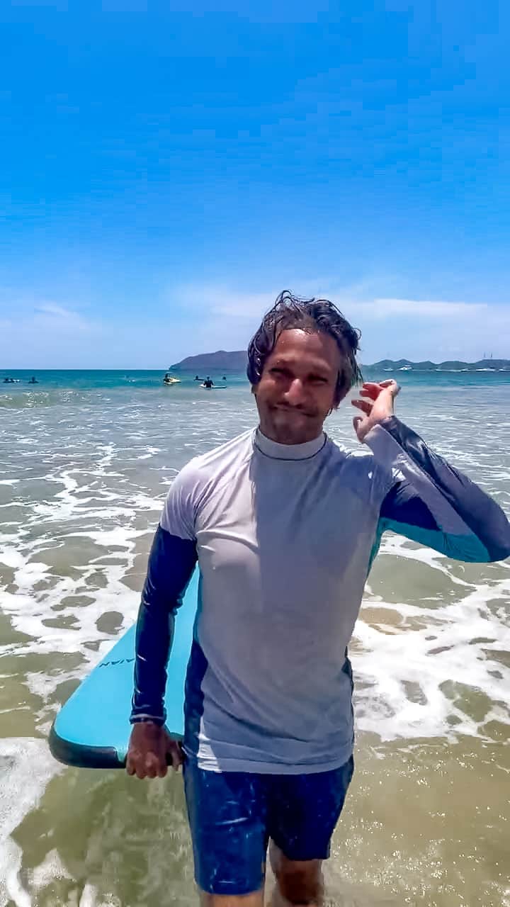 Riz smiling as he walks towards the camera. He is standing in the Pacific Ocean by Tamarindo Beach, and carrying a blue surf board. The sky behind him is clear and a bright blue.