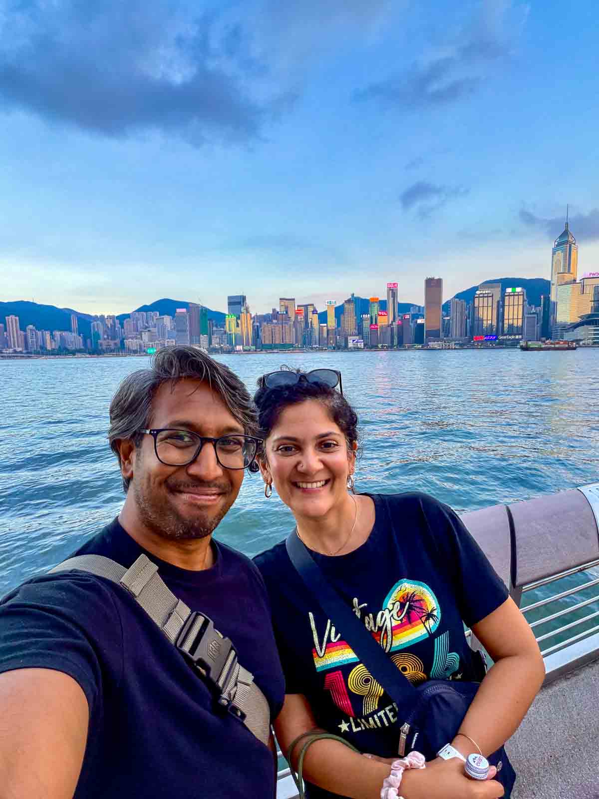 Riz and Reem beside Victoria Harbour at dusk, with illuminated buildings along the Hong Kong Island waterfront.