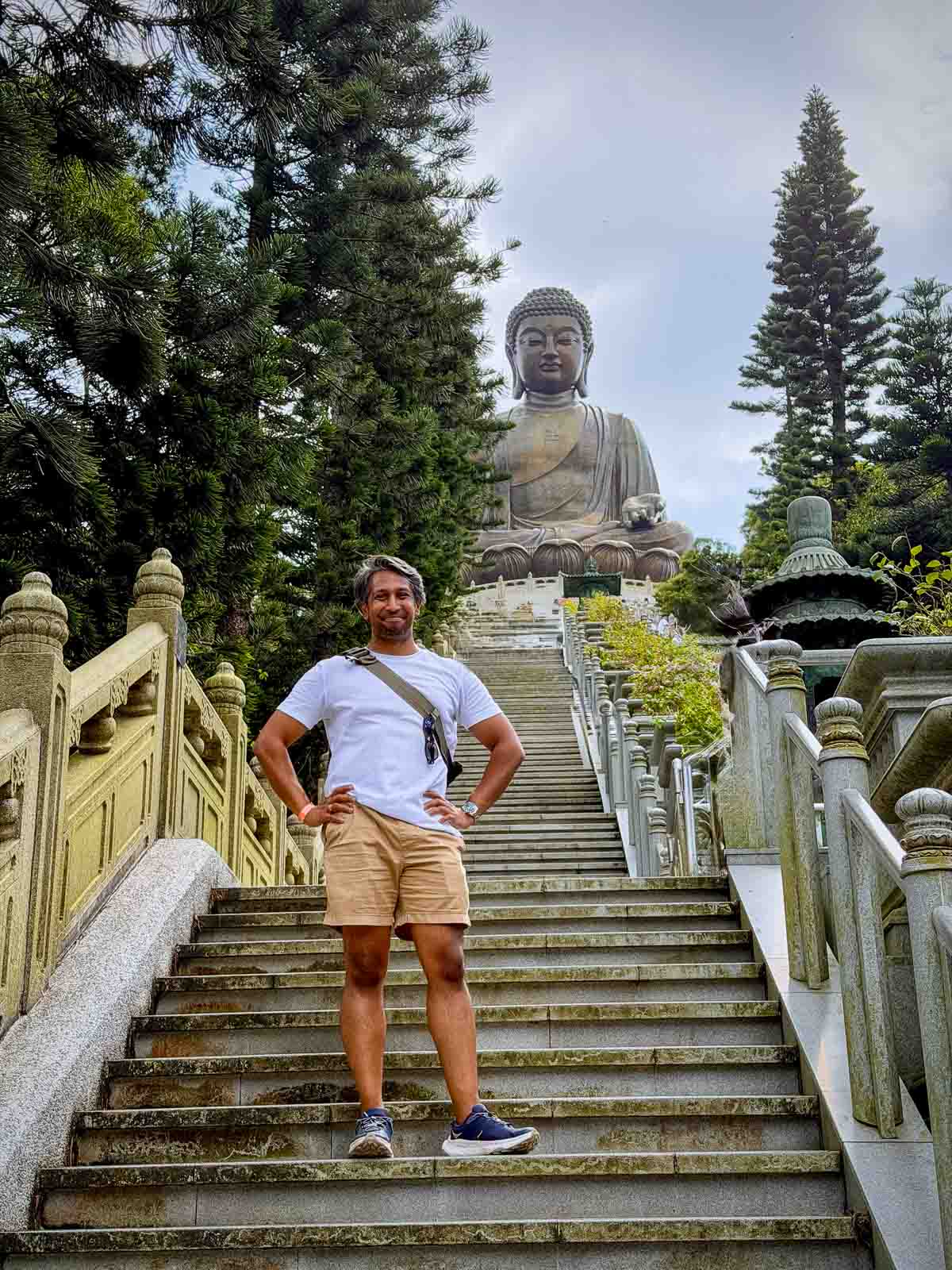 Riz, smiling but visibly posing as a little tired, standing on the stone staircase beneath the Tian Tan Buddha, surrounded by green trees.