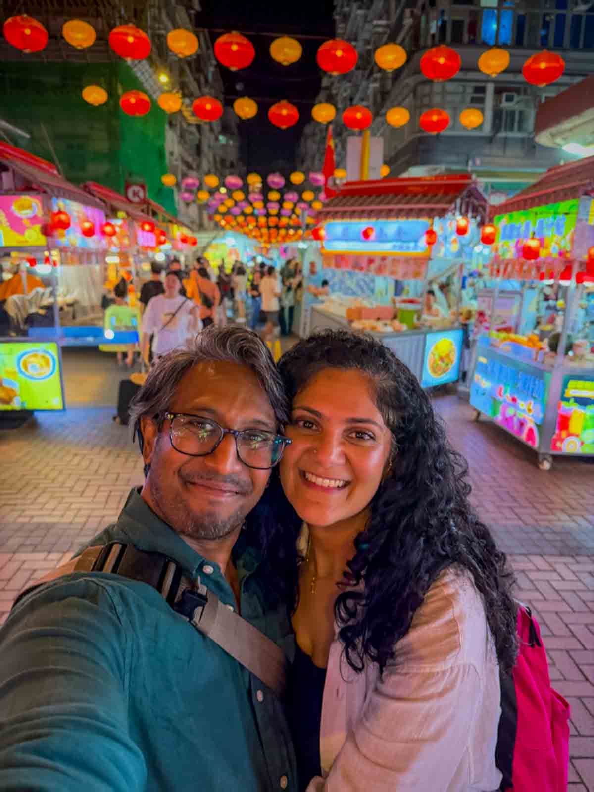 Riz and Reem beneath red and yellow lanterns among the food stalls at Temple Street Night Market.