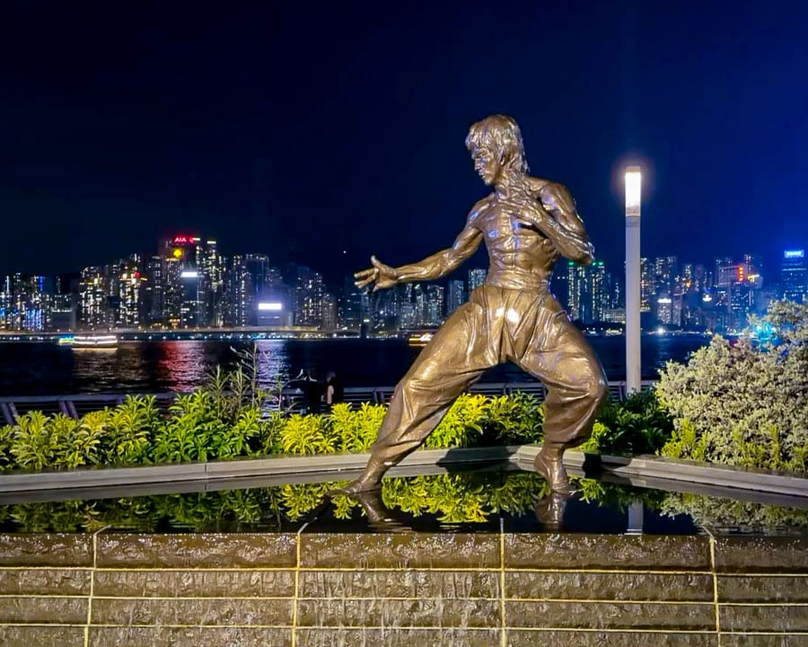 Bruce Lee statue on the Avenue of Stars at night, with Hong Kong's illuminated skyline across Victoria Harbour.