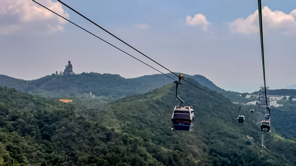 Ngong Ping 360 cable cars above forested hills, with the Tian Tan Buddha visible in the distance.
