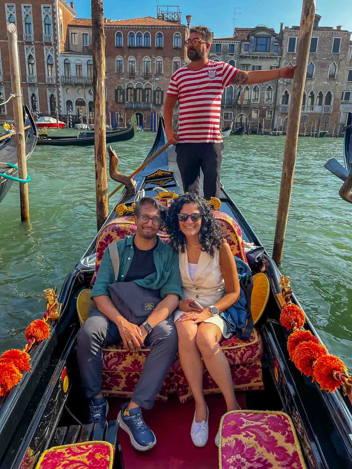Riz and Reem sitting on a colorful gondola in Venice with the traditional gondolier posing behind them.