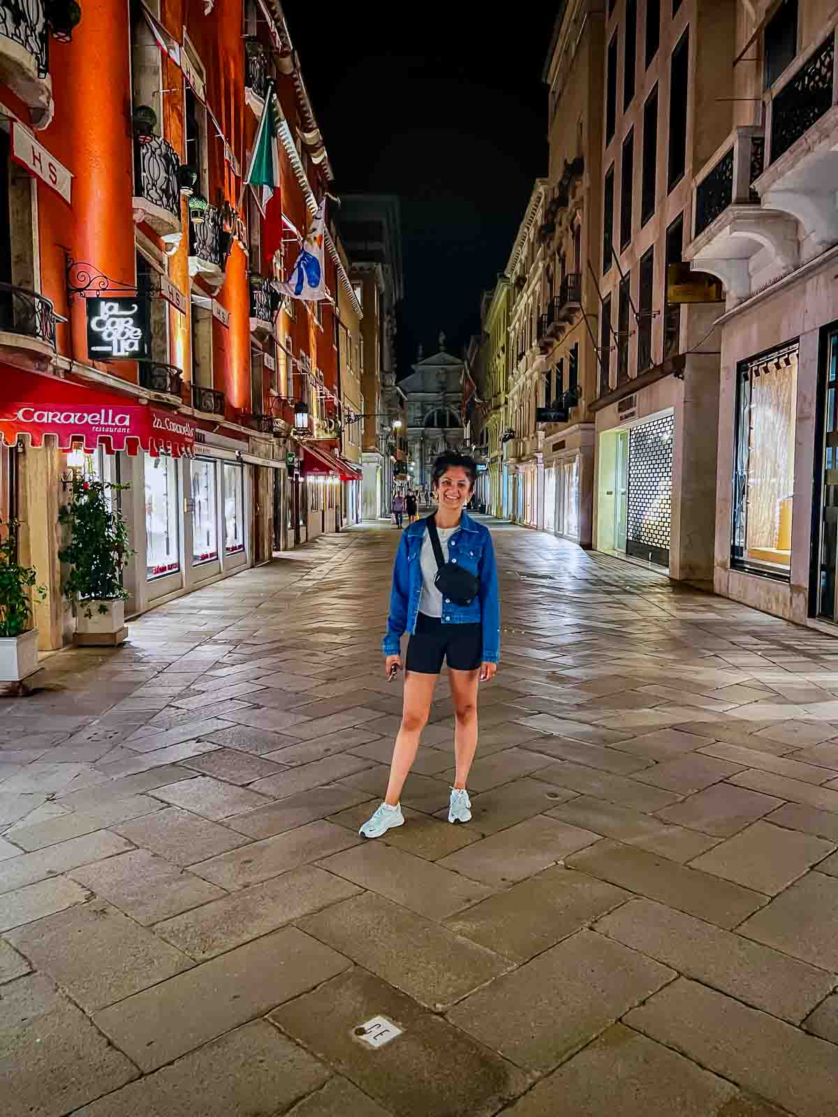 Reem standing alone in a quiet street in Venice at night after the daytime crowds have left. Behind her is La Caravella restaurant.